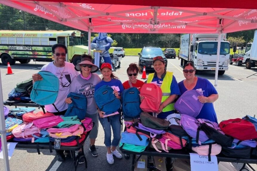 Table of Hope volunteers hold up backpacks at tables stacked with school supplies during a past Backpack Giveaway at County College of Morris.