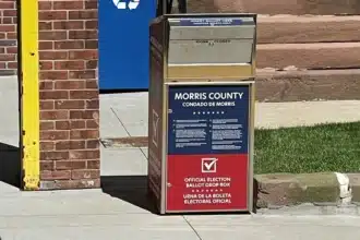 A red, white and blue Morris County official ballot drop box on a sidewalk outside a brick municipal building.