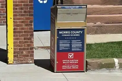 A red, white and blue Morris County official ballot drop box on a sidewalk outside a brick municipal building.