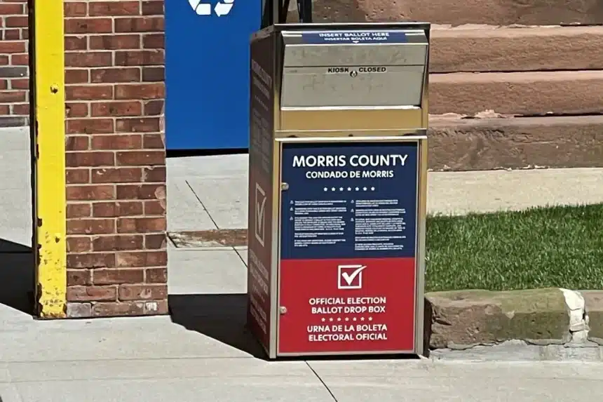A red, white and blue Morris County official ballot drop box on a sidewalk outside a brick municipal building.